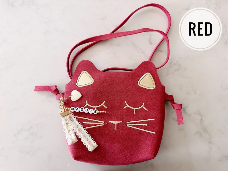 May include: Red cat-shaped crossbody purse with a gold chain and a tassel charm. The charm has a small heart and the name "MADDOIE" spelled out in white beads.