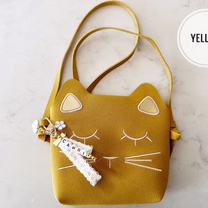 May include: A yellow cat-shaped crossbody bag with a white embroidered face and a gold charm with the name "CAPRI" on it.