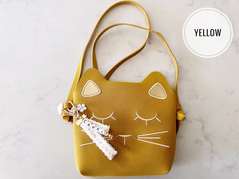 May include: A yellow cat-shaped crossbody bag with a white embroidered face and a gold charm with the name "CAPRI" on it.
