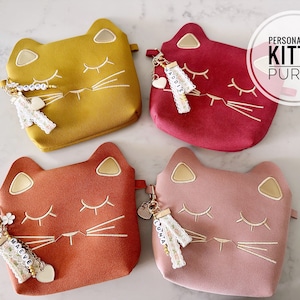 May include: Four personalized kitty purses in different colors: mustard yellow, red, orange, and pink. Each purse has a gold embroidered cat face with closed eyes and whiskers. Each purse has a charm with a name on it: Maddie,  Cuna,  Adddie, and Maeve.