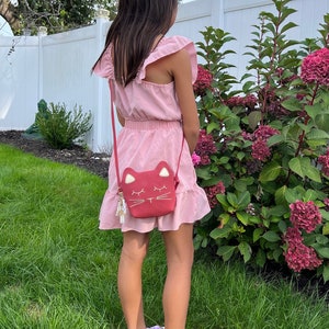 May include: A young person wearing a pink dress with a ruffled hem and a red cat-shaped crossbody bag. The bag has a gold chain strap and a white cat face with closed eyes.