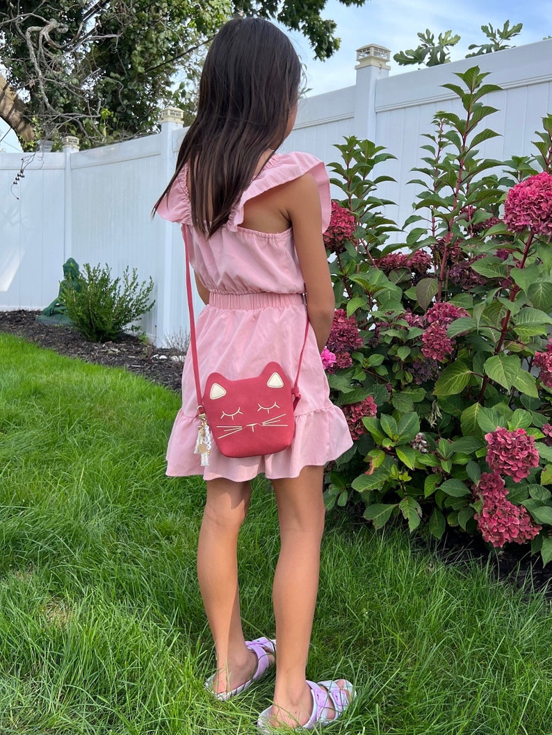 May include: A young person wearing a pink dress with a ruffled hem and a red cat-shaped crossbody bag. The bag has a gold chain strap and a white cat face with closed eyes.
