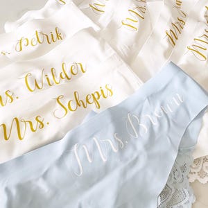 May include: A set of white and light blue lingerie with gold lettering. The lingerie has the words "Mrs. Brown", "Mrs. Wilder", "Mrs. Schepis", "Mrs. Petrik", "Mrs. Smith", and "Mrs. Hughes" written on them.
