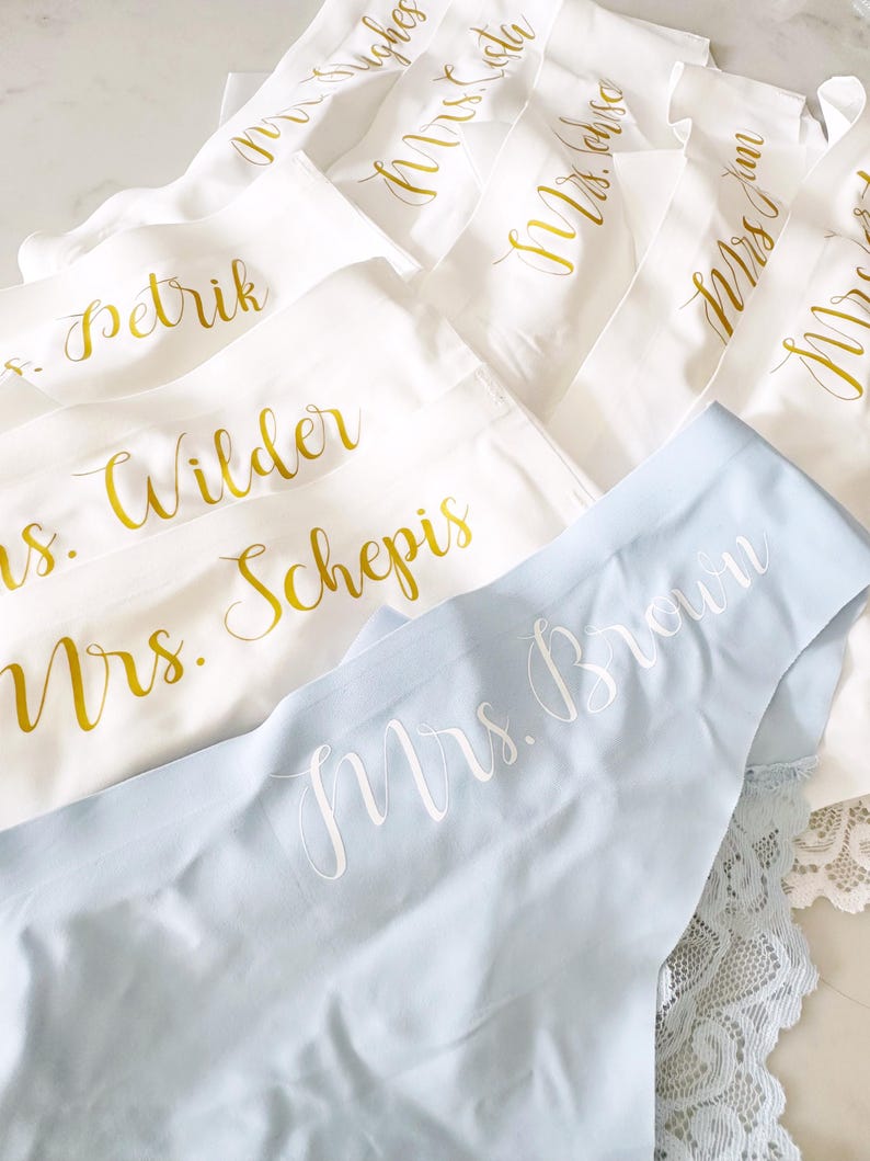 May include: A set of white and light blue lingerie with gold lettering. The lingerie has the words "Mrs. Brown", "Mrs. Wilder", "Mrs. Schepis", "Mrs. Petrik", "Mrs. Smith", and "Mrs. Hughes" written on them.