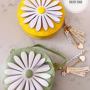 May include: Two daisy-themed bags. One is yellow with a white daisy and a yellow center. The other is light green with a white daisy and a green center. Both have a round shape and a zipper closure. The yellow bag has a charm that says "MAEVE". The green bag has a charm that says "AVA GRACE".