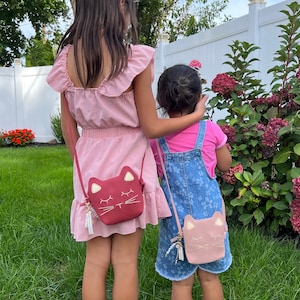May include: Two girls wearing pink and blue dresses with matching cat-shaped purses. The girls are standing in a grassy area with a white fence and a bush with pink flowers in the background.