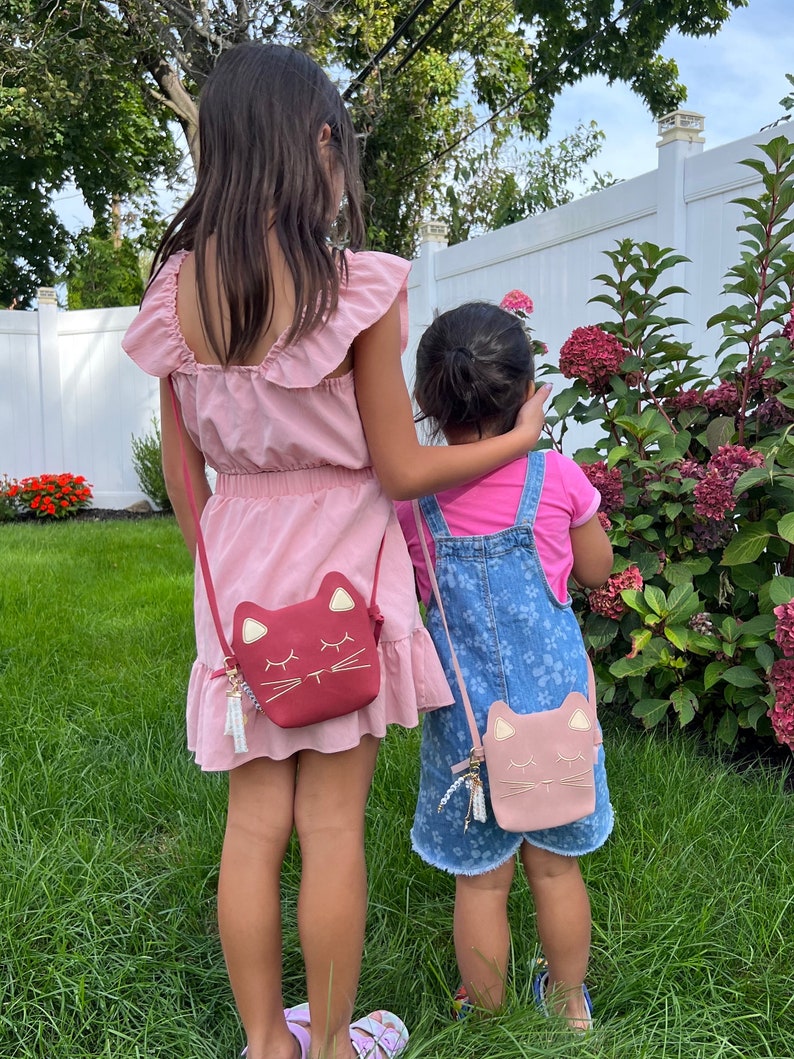 May include: Two girls wearing pink and blue dresses with matching cat-shaped purses. The girls are standing in a grassy area with a white fence and a bush with pink flowers in the background.