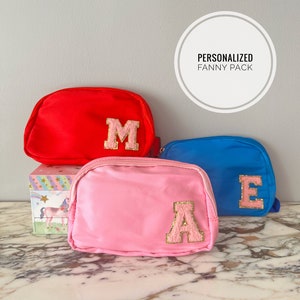 May include: Three personalized fanny packs in red, pink, and blue with gold glitter letters that spell "MAE".  The fanny packs are stacked on top of each other.  The text "PERSONALIZED FANNY PACK" is in a white circle at the top of the image.