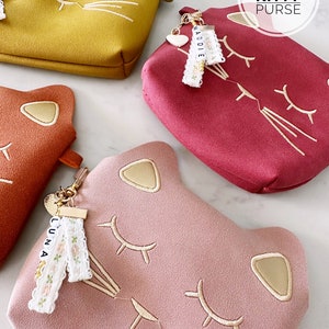 May include: Three pink, red, and yellow kitty-shaped purses with gold embroidery. Each purse has a small charm with a white heart and a name on it. The names are Maddie, Luna, and a third name that is not visible. The purses are on a white surface. The text "Personalized Kitty Purse" is at the top of the image.