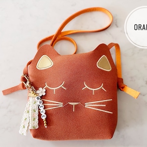 May include: A small orange cat-shaped purse with a gold embroidered face and a white tassel keychain with the name "MAEVE" spelled out in white beads.