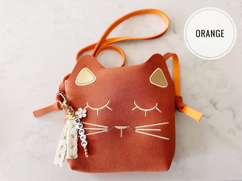 May include: A small orange cat-shaped purse with a gold embroidered face and a white tassel keychain with the name "MAEVE" spelled out in white beads.