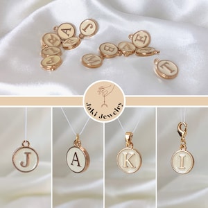 May include: Gold-tone initial charms with white enamel and gold lettering. The letters shown are J, A, K, and I. The charms are on a white background.