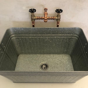 May include: A galvanized metal sink with a copper pipe faucet and black handles. The sink has a drain in the bottom.