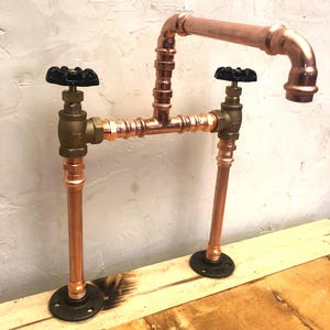 May include: Copper pipe faucet with two black knobs and a copper spout. The faucet is mounted on a wooden surface.