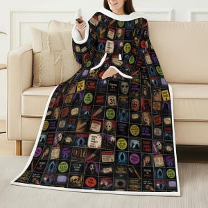 May include: A black hooded blanket with a grid pattern of horror movie posters. The blanket has white trim and a front pocket. The person is holding a remote control. The blanket features text like "STAFF PICKS", "DEATH AWAITS", and "VIDEO NIGHT".