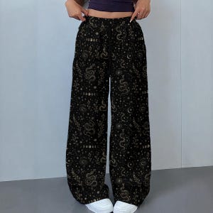 May include: Black wide-leg trousers featuring a repeating gold-toned print of coiled snakes, celestial moon phases, stars, and leafy botanical sprigs. The trousers have a high-rise elastic waistband with a black drawstring cord for an adjustable fit.