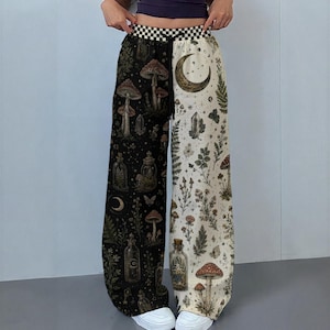 May include: High-waisted wide-leg trousers featuring a split design with a black side and a cream side. The black side displays mushrooms, bottles, and moons, while the cream side features botanical prints, mushrooms, and a crescent moon. A black and white chequered waistband with a black drawstring is included.