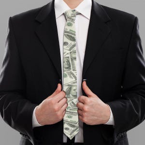 May include: A white tie featuring a pattern of US dollar bills. The tie is being worn by a person in a black suit.