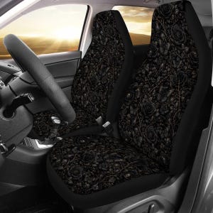 May include: A pair of car seat covers featuring a dark, intricate pattern of black roses, thorny vines, and small skulls set against a deep black background. The covers are fitted over the front seats of a vehicle with a black steering wheel.