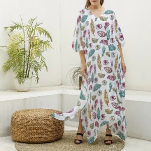 May include: A white floor-length kaftan dress featuring a repeating pattern of colorful feathers and stylized eyes in shades of pink, teal, and yellow. The garment has a V-neckline, loose three-quarter length sleeves, and side slits at the hem.