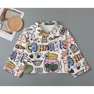 May include: A white button-up jacket with a colorful cartoon print featuring cannabis leaves, marijuana-related words, and other related imagery. The jacket has a button closure and long sleeves.