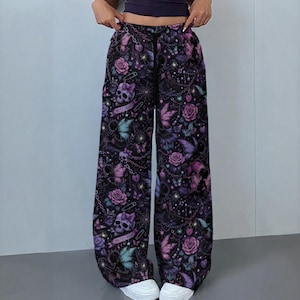 May include: Wide-leg pants in a dark purple and black color palette featuring a repeating pattern of skulls, roses, butterflies, chains, and safety pins. The pants include a black drawstring waistband and a banner with the text Pretty Deadly.