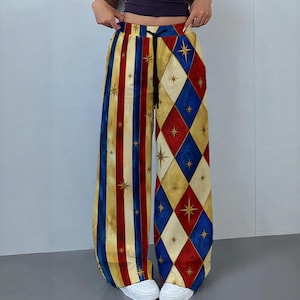 May include: Wide-leg pants featuring a split design with vertical red, blue, and cream stripes on one side and a red, blue, and cream diamond argyle pattern on the other. Both sides feature small gold star accents and a black drawstring waist.