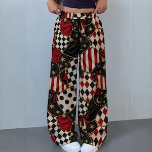 May include: Wide-leg trousers featuring a complex pattern of red, black, and white diamonds, stripes, and polka dots. The design includes gold star and crescent moon motifs, red bows, and gold-colored chain accents with a black drawstring waistband.