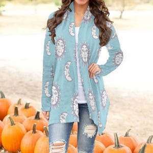 May include: A light blue cardigan featuring a sun and moon design. The long-sleeved cardigan is worn over a white top and paired with ripped blue jeans. Pumpkins are visible in the background.