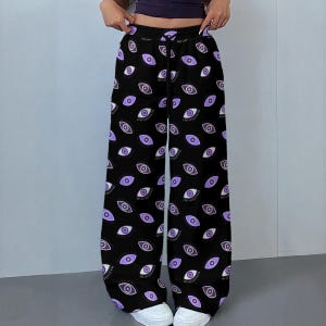 May include: Black wide-leg pants with an all-over eye print in shades of purple and white. The pants have a drawstring waist and are worn with white sneakers. The design features numerous stylized eyes.