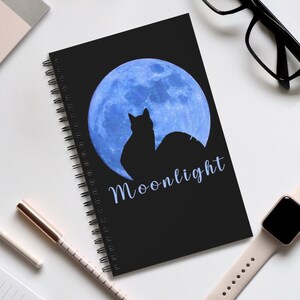 May include: Black spiral bound notebook with a blue moon and a black cat silhouette. The text "Moonlight" is written in white cursive letters.