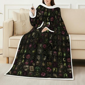 May include: A black hooded blanket with a white trim and pocket. The blanket is covered in a repeating pattern of gothic illustrations in shades of green, pink, and white. The person is sitting on a beige couch.