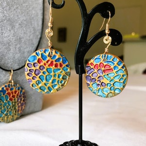 May include: A pair of gold-toned earrings with a blue, red, and purple mosaic design. The earrings are hanging from a black earring stand.