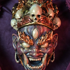 Op de afbeelding: Een gedetailleerd, versierd masker met een demonisch gezicht en een kroon van schedels. Het masker is voornamelijk paars en goud, met witte tanden en schedels. Het ontwerp heeft ingewikkelde details en een dreigende uitdrukking, ideaal voor Halloween of cosplay.