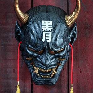 Puede incluir: Una máscara Oni negra y dorada con borlas rojas. La máscara tiene una expresión feroz con dientes y cuernos afilados. La máscara tiene los caracteres japoneses "黒月" escritos en la frente.