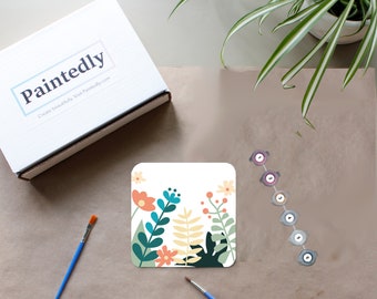 May include: A paint-by-numbers kit with a floral design on a white card. The kit includes seven paint pots with different colors, two paintbrushes, and a box with the text "Paintedly Create beautifully. Visit Paintedly.com".