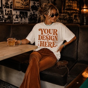May include: An off-white short-sleeve t-shirt featuring the text "YOUR DESIGN HERE" in a bold, orange, retro-style font. The shirt is paired with brown corduroy flare pants, a turquoise beaded bracelet, and dark-tinted sunglasses.