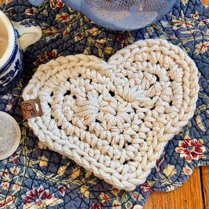 May include: A crocheted heart-shaped coaster in a cream color. The coaster is sitting on a blue and white floral fabric.