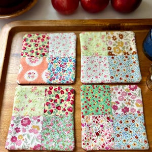 May include: Four square fabric coasters with a patchwork design. Each coaster features a mix of floral and patterned fabrics in shades of pink, green, and blue. The coasters are arranged on a wooden tray, with three red apples in the background.