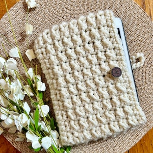 May include: A crocheted tablet case in a cream color with a brown button closure. The case is lying on a beige placemat with a bouquet of white flowers.