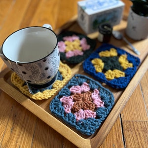 May include: A wooden tray with four crocheted coasters in different colors and patterns. The coasters are yellow, pink, blue, and green. A white mug with a black and white design sits on one of the coasters.
