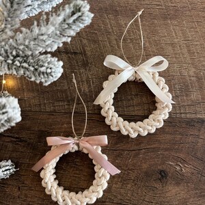 May include: Two macrame wreath ornaments with cream-colored rope, each adorned with a satin ribbon bow. One bow is light pink, the other is cream. The ornaments hang from natural twine loops, perfect for holiday decor.