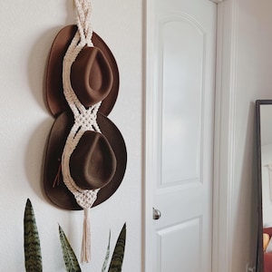 May include: Two brown felt hats hanging on a white macrame hat rack with a tassel. The hat rack is mounted on a white wall.