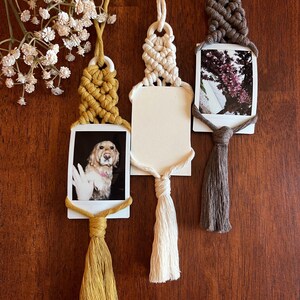 May include: Three macrame photo holders with tassels. The holders are made with white, brown, and yellow cord. Each holder has a small photo attached to it. The photos show a dog, a tree with pink flowers, and a blank white card.