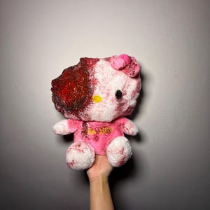May include: A plush toy featuring a white cat character with a pink bow, wearing a pink shirt with the text Hello Kitty. The left side of the head and body displays a textured, dark red, blood-like effect, with similar red splatters across the white fur.