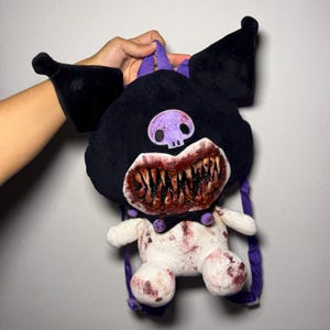 Peut inclure: Un sac à dos en peluche en forme de personnage avec des oreilles pointues noires, un emblème de crâne violet et une large bouche remplie de dents pointues tachées de sang. Le bas du corps est blanc avec des éclaboussures rouges et des bretelles violettes.