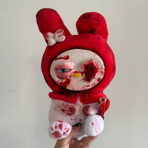 Puede incluir: Un peluche con cara blanca, nariz amarilla, un ojo azul realista y una herida de color rojo oscuro en el otro lado. El personaje lleva una capucha roja brillante con una flor blanca. El cuerpo blanco está cubierto de manchas de color rojo.