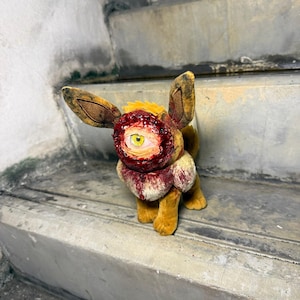 May include: A plush toy resembling a four-legged creature with brown fur, large pointed ears, and a single yellow eye centered in a textured, red, fleshy-looking facial area. The toy features dark, weathered markings and a distressed appearance.