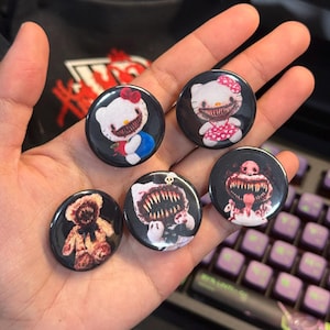 May include: Five circular pin-back buttons featuring dark, horror-themed illustrations on black backgrounds. The designs include two white cat characters with sharp teeth, a teddy bear with a jagged mouth, a skull-topped creature, and a pink monster.