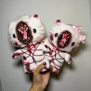 May include: Two white plush cat toys with dark red blood-like stains and red rope bindings. Each toy features a large, open mouth revealing sharp teeth and a pink tongue. One toy has a light pink satin bow attached to the side of its head.